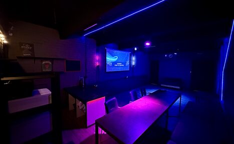 Арендовать Kahovka loft (Каховка Лофт). Неоновый лофт-зал. Тёмное помещение с длинным столом и стульями. Наличие фиолетовой и синей подсветки. На стене экран с изображением. Стеллаж с коробками справа, кирпичная стена слева. Современное оборудование и мебель.