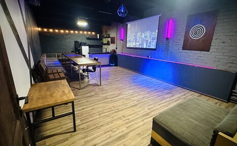 Арендовать Kahovka loft (Каховка Лофт). Неоновый лофт-зал. Пространство с деревянным полом, кирпичными стенами и мебелью из светлого дерева; длинный стол с рядом стульев; диваны вдоль стен; на стене проектор с изображением городской панорамы; разноцветная подсветка по периметру.