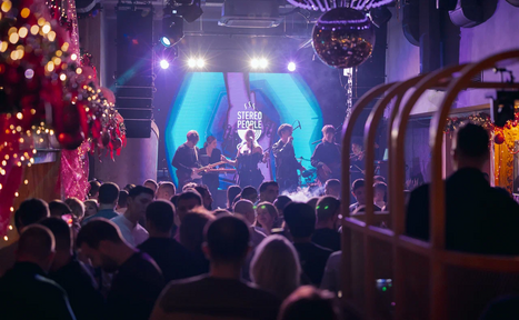 Арендовать Бар StereoPeople (СтереоПипл). Концертное пространство с живой музыкой, большой экран на сцене, группа музыкантов, темное освещение, подвесной светильник в виде дискошара, плотная толпа людей, праздничные декорации с гирляндами и шарами.