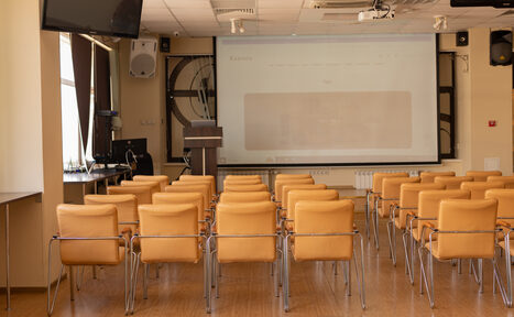 Арендовать Зал 9. Просторный конференц-зал до 200 человек в Академии Кантаты. Конференц-зал с рядами светло-коричневых стульев, деревянный пол, большой экран с проектором, трибунная стойка, светлые стены, потолочные светильники, окна с темными шторами, настенные динамики, кондиционер.