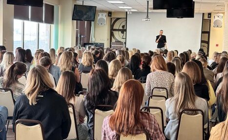 Арендовать Зал 9. Просторный конференц-зал до 200 человек в Академии Кантаты. Конференц-зал с большим количеством людей на стульях, человек в чёрном у белого экрана, микрофоны, две большие колонки на стенах, окна с жалюзи, два телевизора, светлый пол и потолок с люминесцентными лампами.