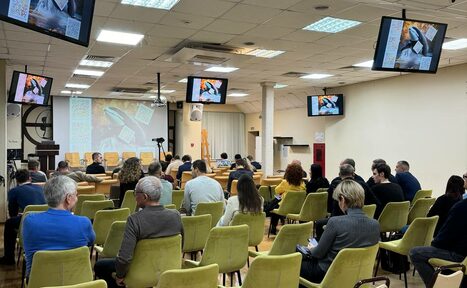 Арендовать Зал 9. Просторный конференц-зал до 200 человек в Академии Кантаты. Конференц-зал с участниками, сидящими на зелёных стульях. Впереди мягкие кресла и проектор, на экранах транслируются изображения. Потолок со встроенным освещением и решетчатыми панелями. На стенах висят экраны. Пол деревянный.