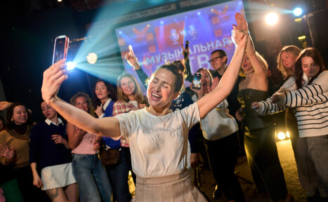 Арендовать Лофт для мероприятий с гольф-симулятором. Человек с телефоном в центре группы людей. Задний план освещён яркими прожекторами, проекция с текстом "Музыкальная" на экране. Несколько человек держат телефоны, создавая эффект мероприятий. Тёмный фон, повседневная одежда.