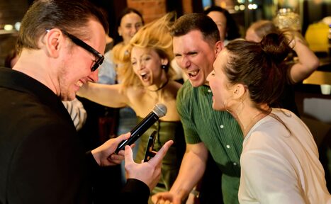 Арендовать Лофт для мероприятий с гольф-симулятором. Изображение показывает группу людей на мероприятии в помещении. Один человек в очках держит микрофон, остальные вокруг в движении, у них радостные выражения лиц. Слева кирпичная стена, видно приглушенное вечернее освещение, барную стойку сзади.