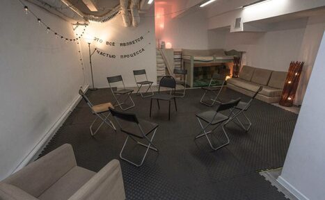 Арендовать Пространство Нора лофт. Комната с черным полом, белыми стенами и мягким освещением. Девять стульев расположены по кругу, слева диван, справа диван и двухъярусная кровать. На стене текст: "ЭТО ВСЕ ЯВЛЯЕТСЯ ЧАСТЬЮ ПРОЦЕССА". Потолок с вентиляционными трубами.