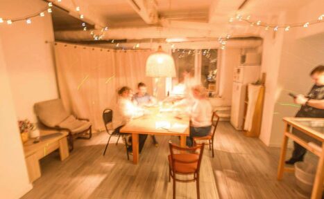 Арендовать Пространство Нора лофт. ```plaintext
Комната с деревянным столом и стульями посередине, мягким креслом слева, холодильником и открытыми полками справа. Светлая плитка на полу, подвесная лампа и гирлянды. Большие окна, зашторенные занавесками. Несколько размытых фигур за столом.
```