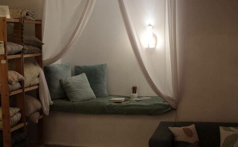 Арендовать Пространство Нора лофт. Пространство с мягкой зоной для отдыха, включающей подушки и книжки, освещённое настенным светильником. Слева деревянные стеллажи с текстилем. Справа диван с подушками. Светлые стены и белый текстиль.