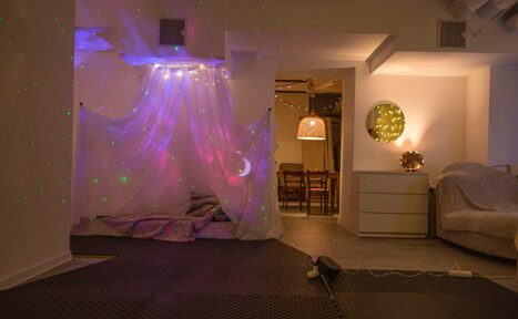 Арендовать Пространство Нора лофт. Комната с зоной отдыха, украшается светящейся занавеской и звездным проектором. Справа комод с круглым зеркалом и лампой. Видны деревянный пол, коврик и стол с деревянными стульями. Мягкое освещение и белые стены.