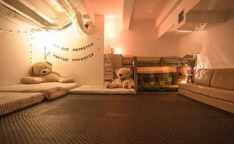 Арендовать Пространство Нора лофт. Подвальное пространство с низкими потолками, ковровое покрытие на полу. На стене текст: "Это все является частью процесса". У стены больших мягких мишек, матрасы, двухъярусная кровать, диван. Мягкое освещение, светодиодные гирлянды.