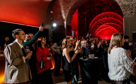 Арендовать Polygon(Полигон). Зал Кухня. Арочный кирпичный зал с приглушенным красным освещением и акцентными светильниками. В центре группа людей стоит вокруг высоких черных столов с напитками. Стены из камня, декорированы светодиодными полосами.