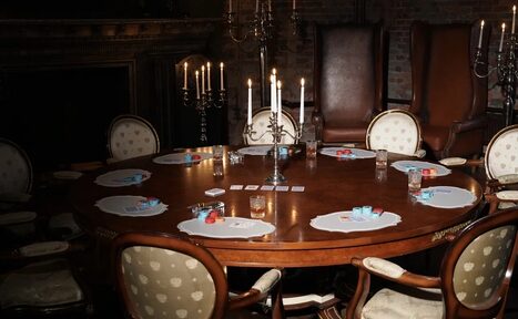 Арендовать Polygon(Полигон). Зал Апартаменты. Круглый деревянный стол с игральными картами и фишками, окруженный стульями с обивкой. На столе свечи в канделябрах. Темное помещение с кирпичными стенами и камином. Тусклое освещение от свечей.