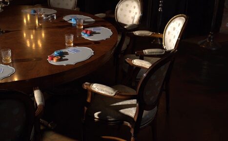 Арендовать Polygon(Полигон). Зал Апартаменты. Круглый деревянный стол с деревянными стульями с обивкой светлого цвета. Несколько стеклянных стаканов, настольные подставки с картами и фишками. Тёмное освещение, тёмный пол, минимум декора.