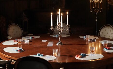 Арендовать Polygon(Полигон). Зал Апартаменты. Круглый деревянный стол с подсвечником по центру, окруженный стульями. На столе представлены подставки, игральные карты и фишки. Обстановка дополняется бокалами и свечами на подсвечниках, обеспечивающими мягкое освещение. Цвета темные, детали классические.