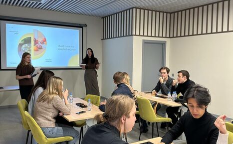 Арендовать Зал 6. Просторный и светлый конференц-зал до 50 человек в Академии Кантаты. Современное офисное пространство с людьми за столами. Стены светлые, потолок с решеткой. Мебель: небольшие деревянные столы, желтые стулья. Зона презентации с проектором и экраном. На экране текст и изображения. Все присутствующие заняты обсуждением.