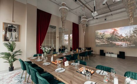 Арендовать Rubin Loft (Рубин Лофт). Зал Олд Мани. Просторное помещение с высоким потолком и открытыми коммуникациями. Длинный деревянный стол с зелеными стульями. Красные шторы, большие зеркала, проектор с изображением пейзажа, декоративные элементы, люстры, растения. Светлые стены, паркетный пол.