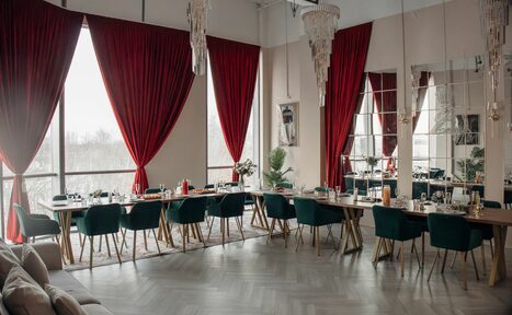 Арендовать Rubin Loft (Рубин Лофт). Зал Олд Мани. Просторное помещение с большими окнами, закрытыми красными шторами. Длинные столы с сервировкой, вокруг темно-зеленые стулья. На полу паркет светлого оттенка. Потолочные хрустальные люстры. Белые стены, на них большие зеркала.