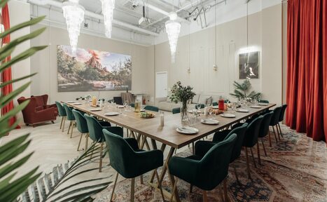 Арендовать Rubin Loft (Рубин Лофт). Зал Олд Мани. Просторная комната с длинным деревянным столом и зелеными стульями на металлических ножках. На столе посуда и напитки. Светильники с подвесными кристаллами. Окружение дополнено красными шторами, картинами, ковром и живыми растениями. Светлые стены и высокий потолок.