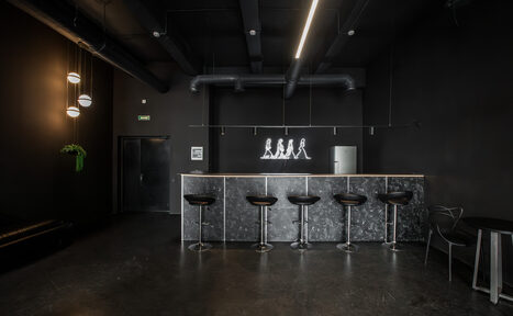 Арендовать Rubin Loft (Рубин Лофт). Лофт Блэк. Барная стойка с пятью высокими табуретами, черные стены и потолок с открытыми коммуникациями, неоновая инсталляция, подвесные светильники, металлический холодильник, выходной знак, черный диван.