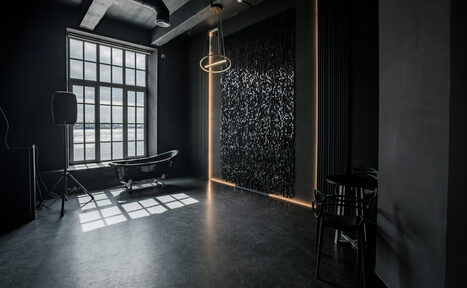 Арендовать Rubin Loft (Рубин Лофт). Лофт Блэк. Лофт-пространство с высоким потолком, большим окном, вид на улицу. Чугунная ванна у окна. Черные стены с текстурной панелью и светодиодной подсветкой. Металлическая подвесная лампа. Три стула у стены. Пол темный, бетонный. Освещение естественное и искусственное.