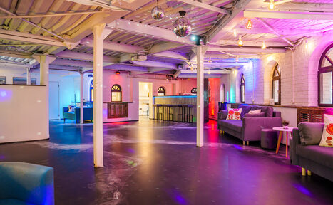 Арендовать Лофт с террасой на территории Измайловского Вернисажа. Просторное помещение с открытой планировкой, включая барную стойку с высокими стульями. Мягкие диваны серых тонов, деревянные столики. Потолок с открытыми балками и разноцветная подсветка. Диско-шары и гирлянды. Белые стены с арочными окнами. Черный пол.
