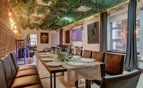 Арендовать Лофт Семеновский. Пространство с кирпичной стеной и подвесным декором из зелени. Длинный обеденный стол с сервировкой на восемь человек, кожаные стулья. Окна с темными шторами. Потолочные светильники, светлые плиточные полы, проектор и экран.