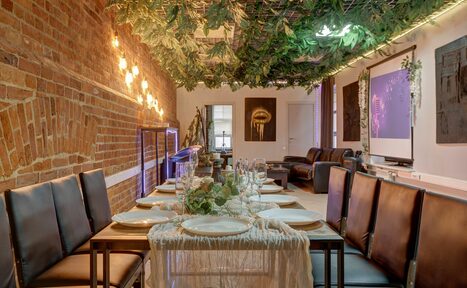 Арендовать Лофт Семеновский. Длинный стол с сервировкой и черными стульями, кирпичная стена слева, потолок украшен зелеными растениями, неоновая подсветка, на стене картины и проектор, сзади диваны, оформлено в теплых тонах, есть декоративные растения.