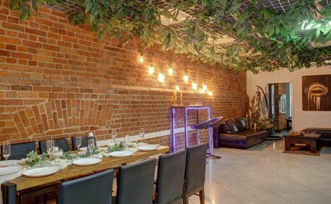 Арендовать Лофт Семеновский. Ресторанное пространство с кирпичными стенами и подвесной декорацией из зелени. Длинный стол, сервированный тарелками и бокалами, с черными стульями. Мягкий диван в углу. Настенные светильники, картина. Пол выложен плиткой.