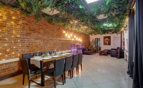 Арендовать Лофт Семеновский. Просторное помещение с кирпичной стеной, вдоль которой стоит накрытый стол и десять стульев. Потолок украшен зеленью, пол выложен светлой плиткой. Освещение представлено гирляндами и лампами. В дальней части виден кожаный диван.