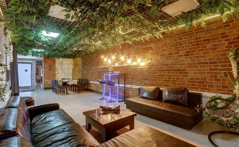 Арендовать Лофт Семеновский. Лофт-пространство с кирпичными стенами. Диваны и столы из темного дерева. Потолок с зелеными декоративными растениями. Подвесные светильники на стене. Плиточный пол светлого цвета. Стойка с подсветкой. Планировка с зонами для отдыха и приема пищи.
