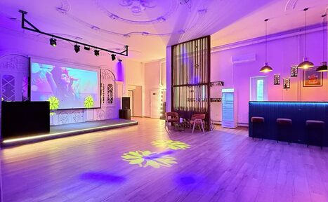 Арендовать Лофт Софт. Просторное помещение с деревянным светлым полом, сценой, подсветкой фиолетового и желтого цветов. Справа — барная стойка с высокими стульями. На стене — большой экран с проектором. Потолок украшен лепниной, подвесные лампы над баром.