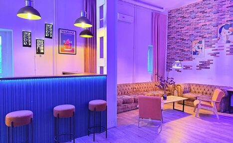 Арендовать Лофт Софт. Лофт-пространство с барной стойкой и тремя стульями, мягкий диван, кресло и стол. Яркое освещение, разноцветные неоновые акценты, кирпичная стена с картинами, светлый деревянный пол, фиолетовые шторы. Настенные светильники и подсветка стойки.