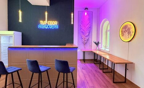 Арендовать Лофт Лайф. Барная зона с высокими стульями, неоновая надпись на тёмной стене, круглое настенное свечение, длинные деревянные столы вдоль стены, декоративные элементы, подвесные светильники, арочное окно, деревянный пол.