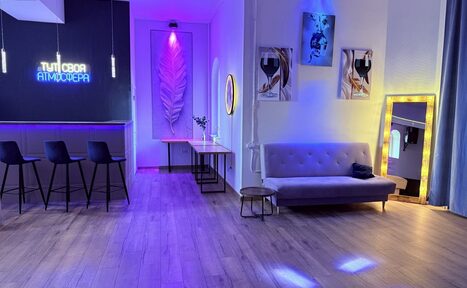 Арендовать Лофт Лайф. Барное пространство с деревянным полом, фиолетовой и синей подсветкой. Стойка с четырьмя черными барными стульями. Диван, два постера, зеркало с подсветкой. Неоновая надпись на стене, деревянный стол с цветами.