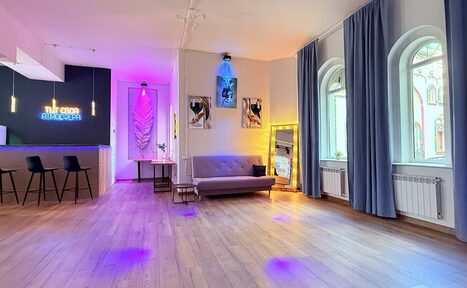 Арендовать Лофт Лайф. Просторное помещение с деревянным полом, бежевыми стенами и арочными окнами с синими занавесками. Барная стойка с двумя стульями слева. Диван, зеркало и картина на правой стене. Разноцветная подсветка и неоновая надпись на черной стене.