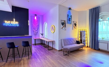 Арендовать Лофт Лайф. Современное пространство с барной зоной и трех стульями, диван, зеркалo, столы. Неоновая подсветка, окна с арками, паркетный пол. На стене постеры, надпись на стене "Тут своя атмосфера". Занавески слева, радиатор под окном.
