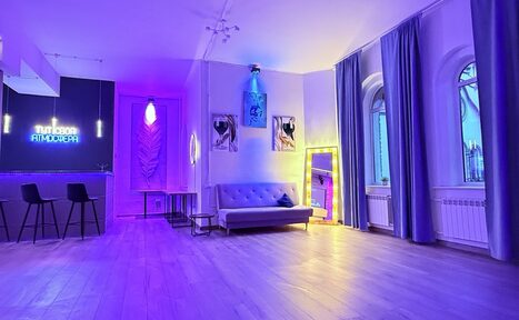 Арендовать Лофт Лайф. Просторное помещение с диваном и зеркалом, украшенное неоновыми вывесками и картинами. Барная стойка с двумя стульями. Большие окна с занавесками, деревянный пол, фиолетовое и жёлтое освещение.