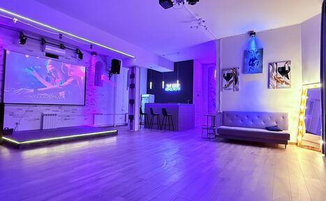 Арендовать Лофт Лайф. Просторное помещение с деревянным полом, сцена с экраном, барная стойка с высокими стульями, серая софа, неоновая подсветка, стены с постерами и подсветкой, большое зеркало в раме с лампочками, фиолетовое и синее освещение.