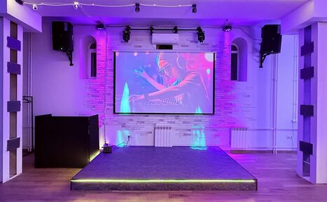 Арендовать Лофт Лайф. Пространство с небольшой сценой, оборудованной экраном и колонками. Потолок с трековыми светильниками, стены белые с отделкой под кирпич, деревянный пол. Сцена подсвечена разноцветным светом, установлен диджейский пульт.