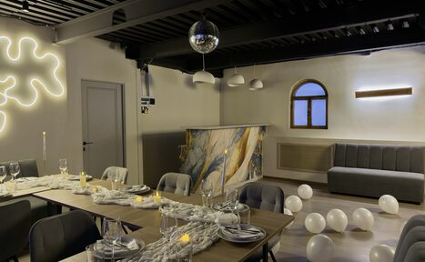 Арендовать Maybe Loft (Мэйби Лофт). Зал Olive (Олив). Современный дизайнерский лофт . Пространство для мероприятий с длинным деревянным столом, сервированным стеклянной посудой и свечами. Мягкие серые стулья, изогнутая серая скамья, стойка с мраморным узором, диско-шар, подвесные светильники, белые шары на полу, декоративная подсветка на стене.