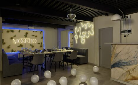 Арендовать Maybe Loft (Мэйби Лофт). Зал Olive (Олив). Современный дизайнерский лофт . Современное пространство с длинным столом и серыми стульями, на столе накрыты приборы и свечи. Стены украшены неоновыми знаками и элементами. Потолок черный, на полу белые шары. Барная стойка с мраморной текстурой и декоративное освещение.
