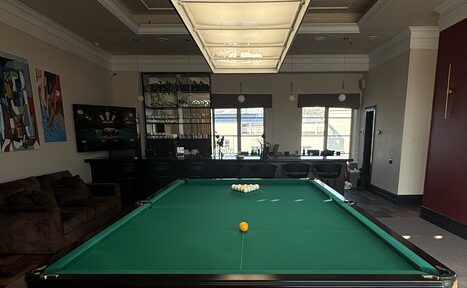 Арендовать Студия Н. Бильярдная комната с зеленым бильярдным столом в центре. На заднем плане барная стойка с высокими стульями и полки с бокалами. На стене висят картины. Освещение: потолочный светильник над столом. Цвета: темные и нейтральные.