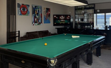 Арендовать Студия Н. Бильярдная с зелёным столом и шарами, рядом диван, на стенах картины. В углу стеллаж с бутылками, бокалом и телевизором. Светлый потолок с подвесным светильником. Холодный свет от окна. Светлые стены, ковровое покрытие пола.