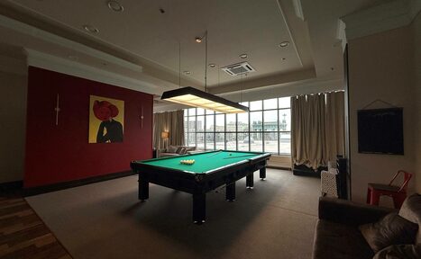 Арендовать Студия Н. Просторная комната с бильярдным столом в центре, красная стена с картинами, большая панорамная витражная окно с занавесками, мягкий диван и кресло, подвесной светильник над столом, паркетный пол.