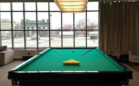 Арендовать Студия Н. Бильярдная комната с большим столом, зеленым сукном, двумя киями и набором желтых шаров. За столом панорамные окна с видом на улицу. Слева диван, справа кресло с узором. Потолочный светильник, бежевые шторы.