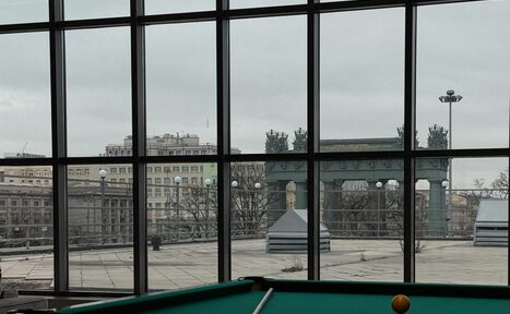 Арендовать Студия Н. Пространство с большими панорамными окнами, за которыми видна терраса и городская архитектура. Внизу части окна расположен стол для бильярда с зеленым сукном и шаром. Потолочный светильник не включен. Цвета: серый, зеленый.