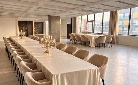 Арендовать Loft Prostranstvo Huge(Огромное Лофт Пространство). Просторное помещение с большими окнами, длинный стол с белой скатертью и золотыми канделябрами, мягкие стулья бежевого цвета, пол из светлого дерева или ламината, потолок с балками, светлые стены, много естественного света.