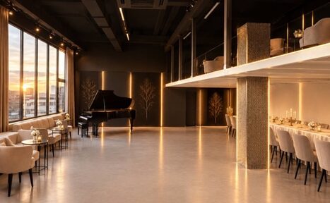 Арендовать Loft Prostranstvo Huge(Огромное Лофт Пространство). Просторная комната с высоким потолком, фортепиано слева, столики с креслами вдоль панорамных окон, справа длинный стол с сервировкой на полуподнятой платформе. Черные стены с декоративными элементами, теплое освещение.