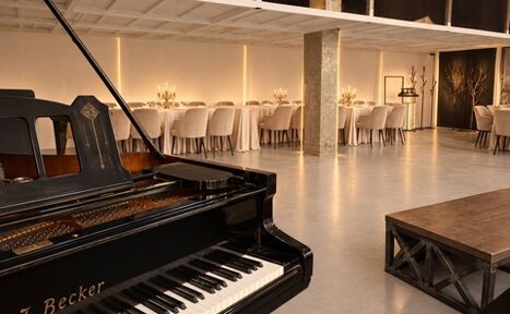 Арендовать Loft Prostranstvo Huge(Огромное Лофт Пространство). Помещение со столами и стульями, украшенными скатертями и чехлами, в центре черный рояль, справа деревянный стол. Стены светлые с мягкой подсветкой. Пол бетонный. Открытая планировка, высокие потолки.