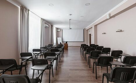 Арендовать Гостиница Сокол. Конференц-зал. Конференц-зал с рядами черных стульев с откидными столиками, проекторным экраном у передней стены, деревянным напольным покрытием, бежевыми стенами и белым потолочным освещением. На стене висит белая доска. Возле окна – шторы.