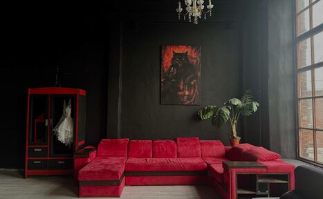 Арендовать C. A. T. Group (C.A.T. Группа). Пpoстоpный двуxэтaжный лoфт. Комната с черными стенами и открытым потолком. Красный угловой диван, стеклянный шкаф с белым платьем, картина на стене, крупное растение в горшке. Большое окно с металлическими переплетами справа, подвесная люстра. Тёмное дерево и текстура.